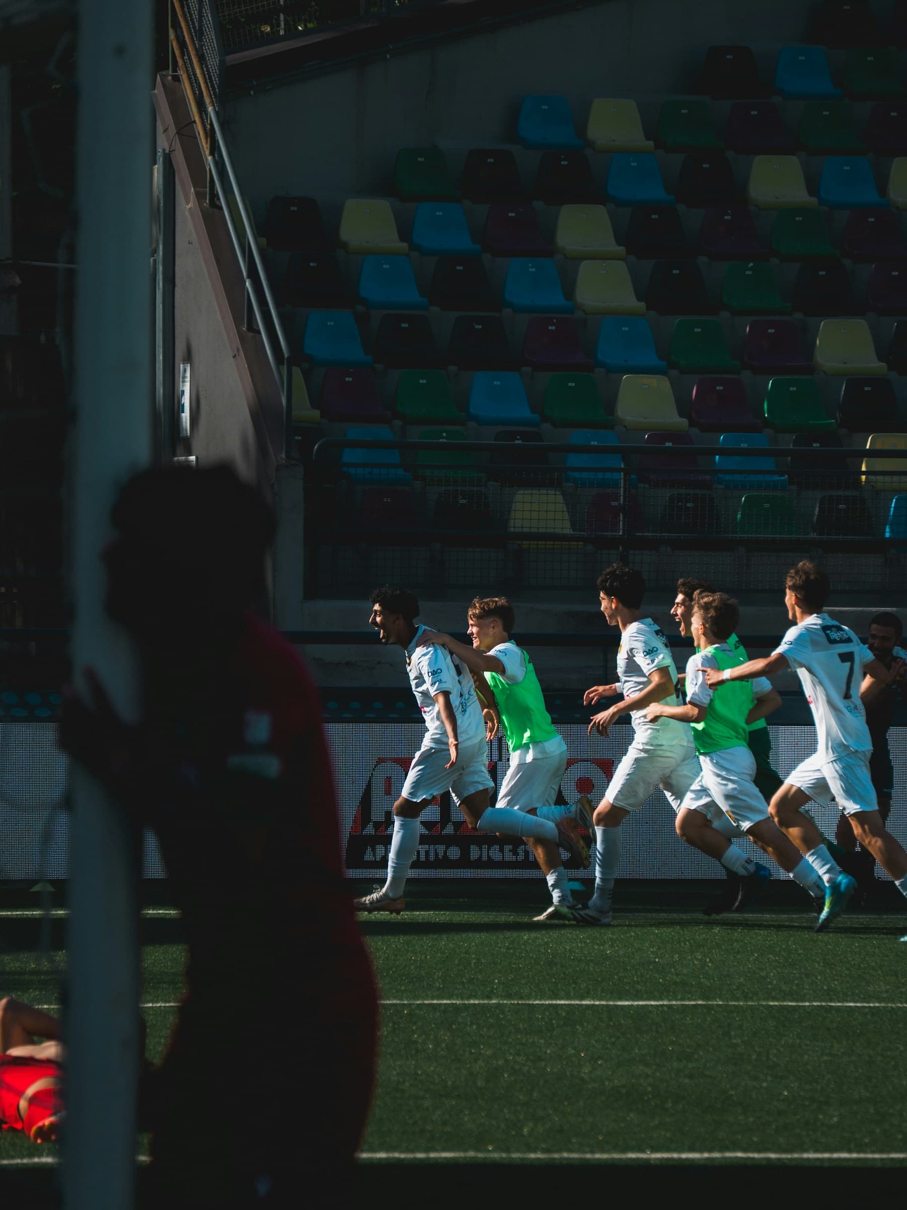 Footballeurs en blanc qui courent fêter un but, devant une tribune aux sièges colorés